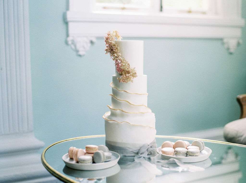 White three tier cake with gold wrapped ruffles and silk grey ribbon on a glass table with two plates of blush and grey macarons