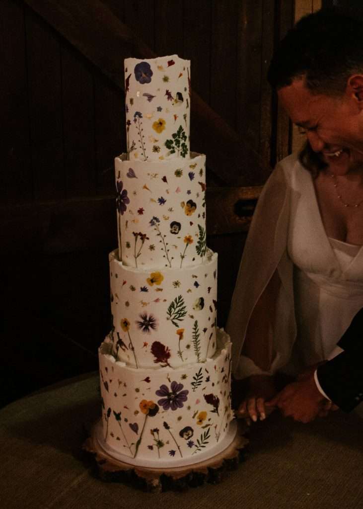 Couple cutting a tall four tier buttercream cake with fresh flowers in a barn
