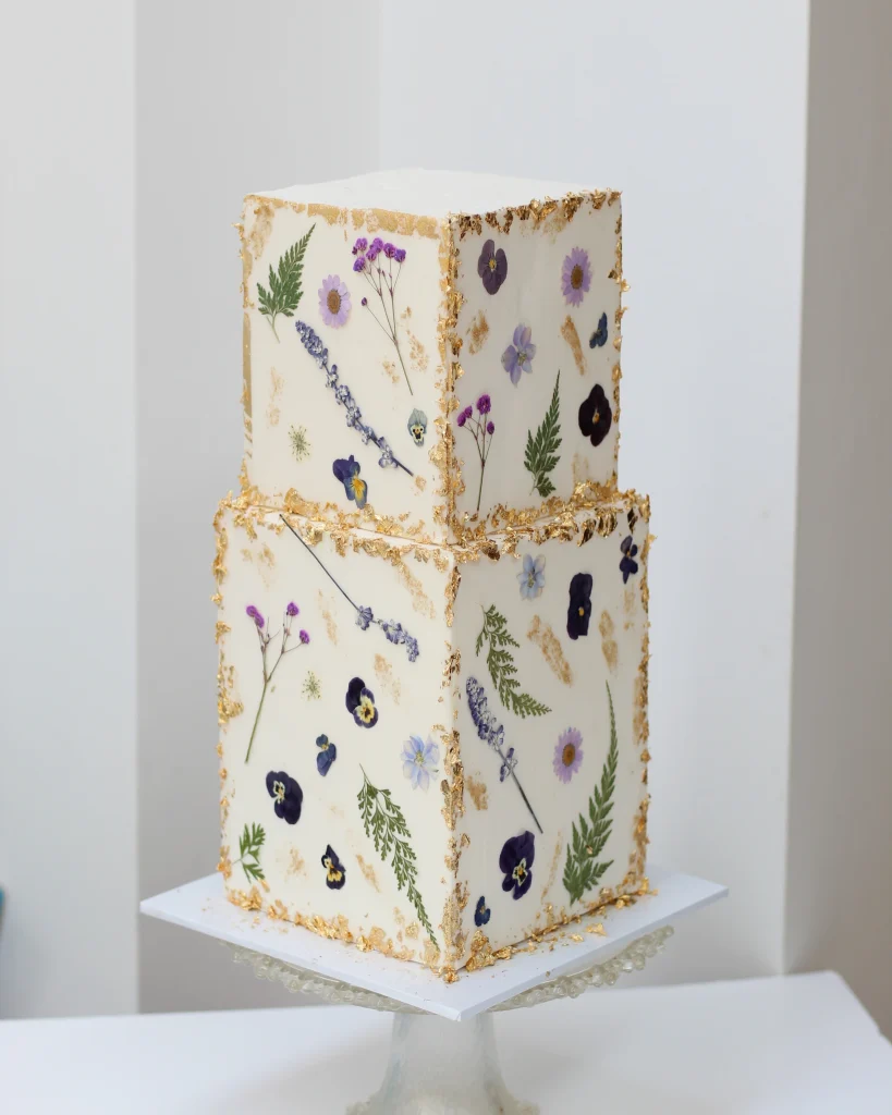 White two tier square wedding cake with multi coloured pressed flowers and gold leaf